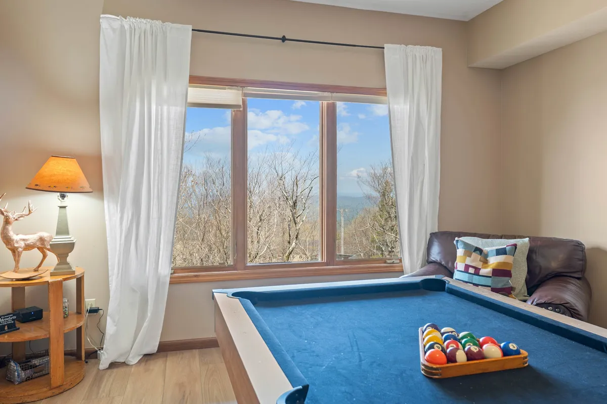 This interior image features a pool table, comfortable seating, large windows with expansive outdoor views, and a decorative lamp. The space is designed for casual gathering and entertainment, while the generous windows frame the colorful seasonal landscape outside.