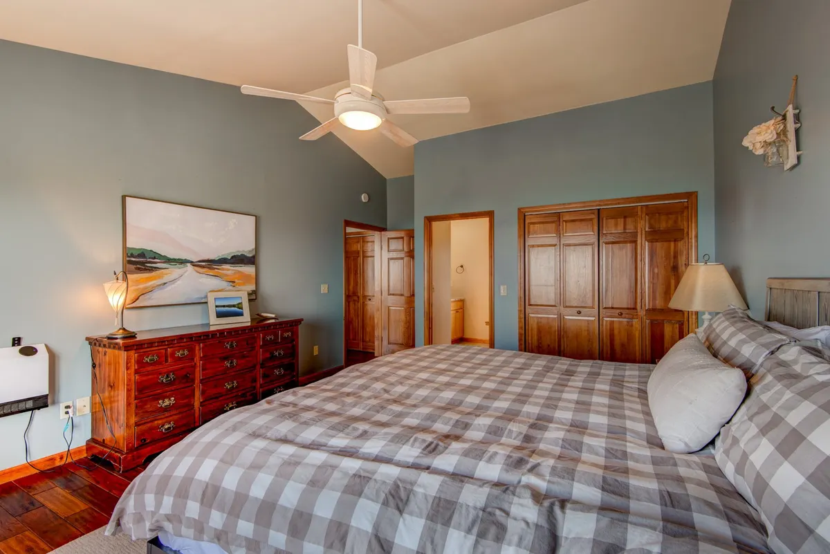 A serene bedroom featuring a large bed with a plaid comforter, a wooden dresser, a framed landscape painting, ceiling fan, and wooden closet doors. Soft lighting and neutral tones create a cozy, restful atmosphere ideal for relaxation after a long day.