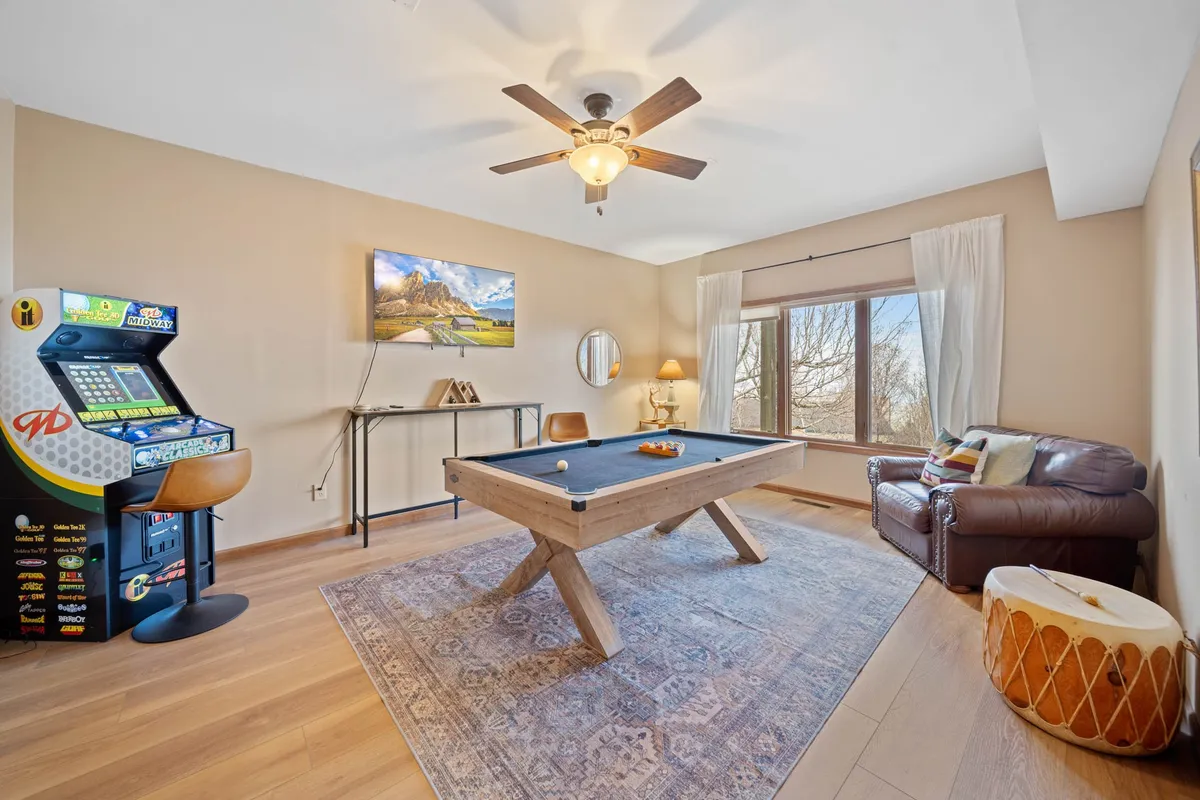 A lively game room atmosphere with a pool table, arcade game, leather sofa, large window, and a TV displaying landscape art. Natural light pours in, hinting at the inviting outdoors just beyond.