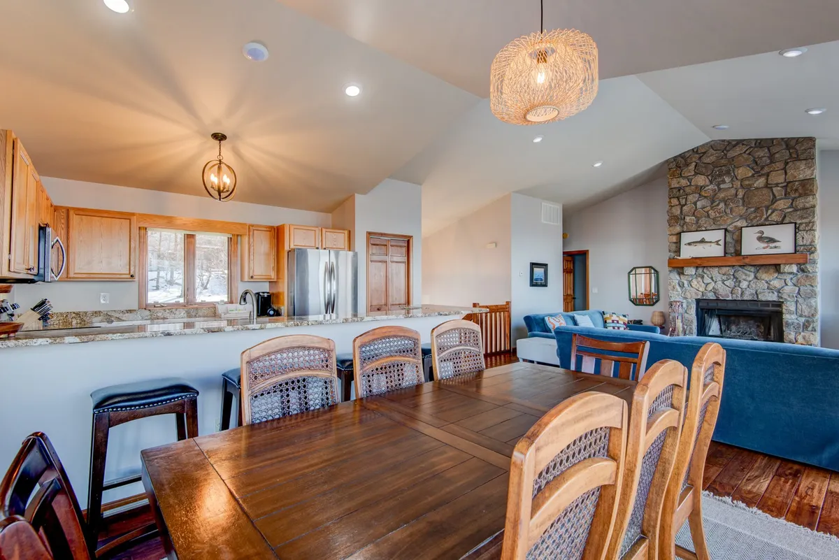 A spacious open-plan area featuring a wooden dining table adjacent to the kitchen, a cozy fireplace, and a plush sofa, all set in a warmly lit environment. The layout encourages easy movement between cooking, dining, and relaxing, fostering togetherness.