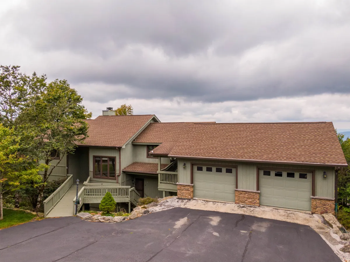 This image showcases the home's welcoming facade, attached garage, and surrounding landscaping, all bathed in warm light. The front porch is styled for relaxation and gathering, suggesting an inviting entry point for guests coming to enjoy the mountain surroundings and attend local festivities.