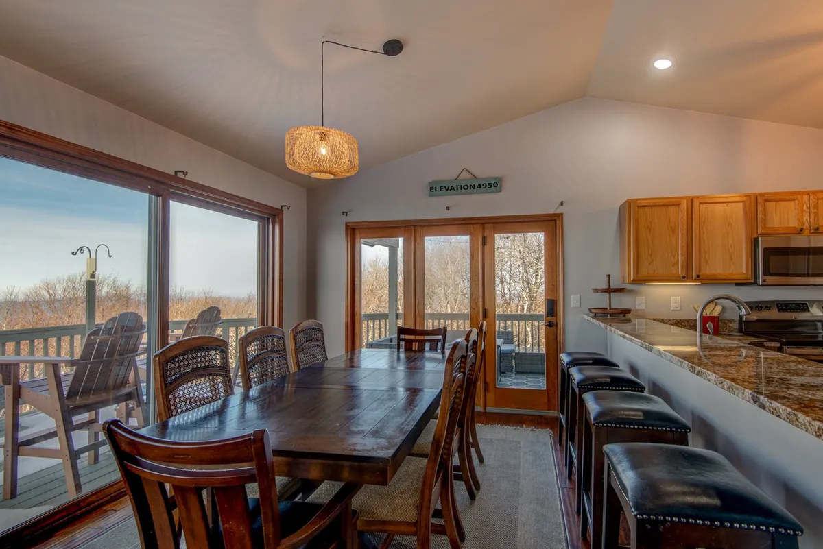The image spotlights a large wooden dining table with ample seating and large windows that offer a stunning scenic view. The kitchen bar area is visible, supporting the idea of everyone staying connected during meals and conversations. Natural light and thoughtful decor create a warm, communal mood.