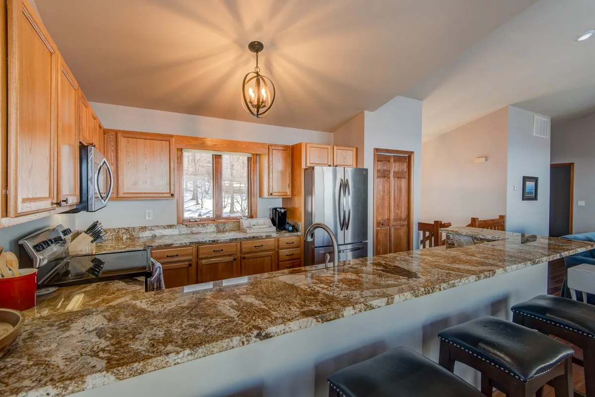 Close-up of a granite kitchen countertop, wood cabinets, inviting light fixtures, and bar stools lining the breakfast bar. The scene focuses on the functional and social aspects of the kitchen, emphasizing a cozy spot for guests to chat or enjoy a meal while cooking takes place.