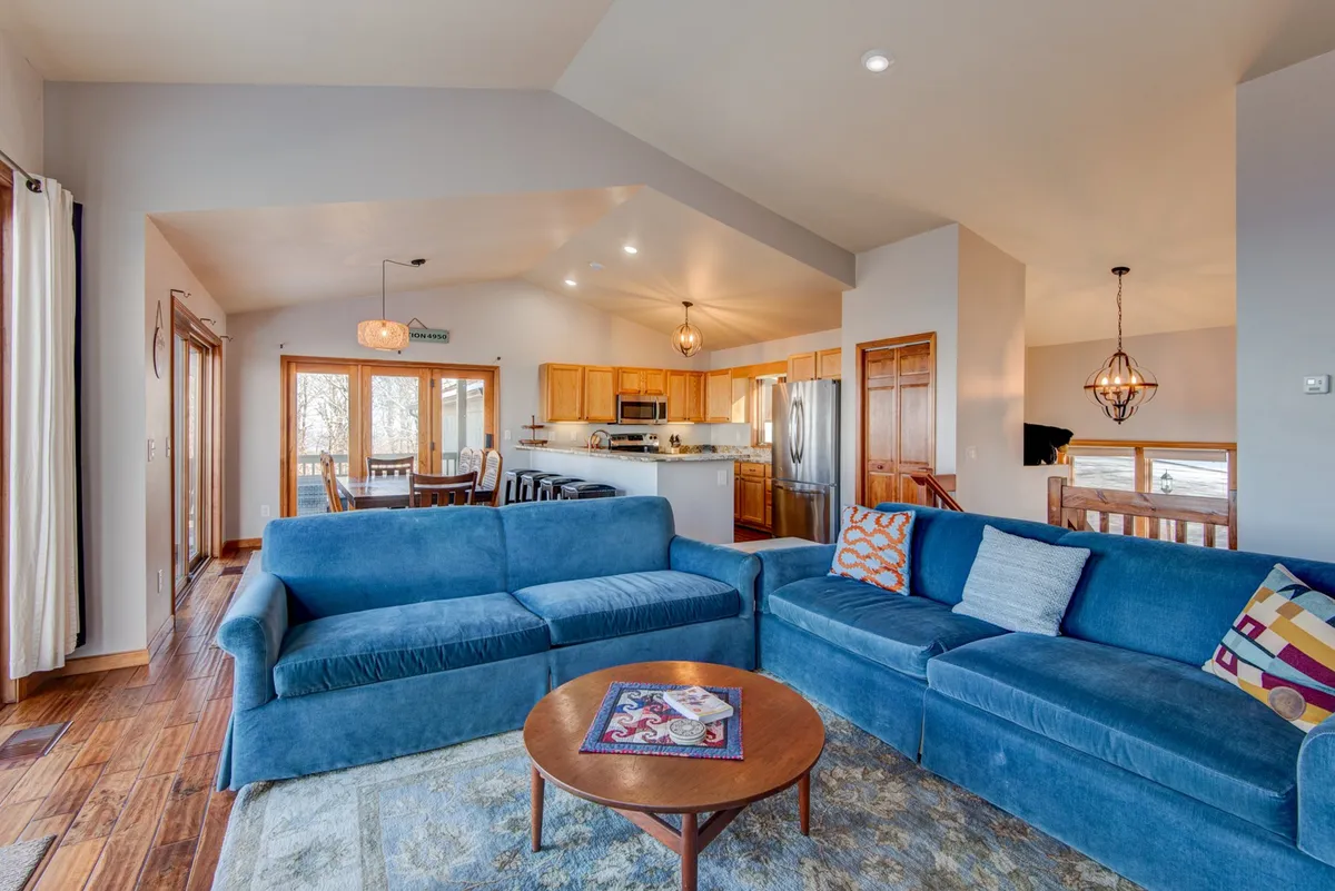 This image captures the flow between the living area, wooden dining table, and nearby kitchen. The blue couch, decorative rug, and wood accents create a cohesive, welcoming space ideal for socializing and family gatherings. The dining area is seamlessly integrated, reinforcing the open-plan layout described in the section.