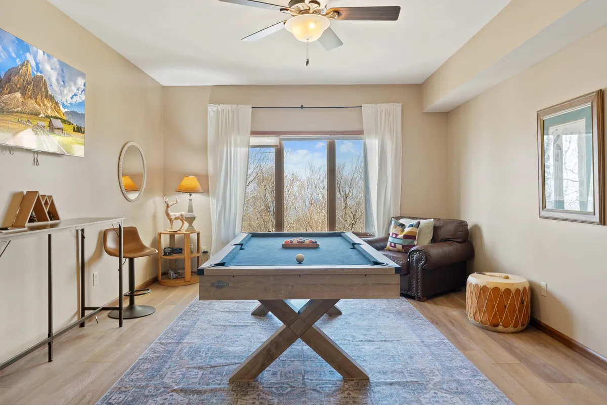 A spacious, bright recreation room featuring a pool table, comfortable seating area, large windows letting in natural light, and decorative artwork. The view through the windows hints at the surrounding outdoor scenery, connecting the interior space with the mountain landscape outside.