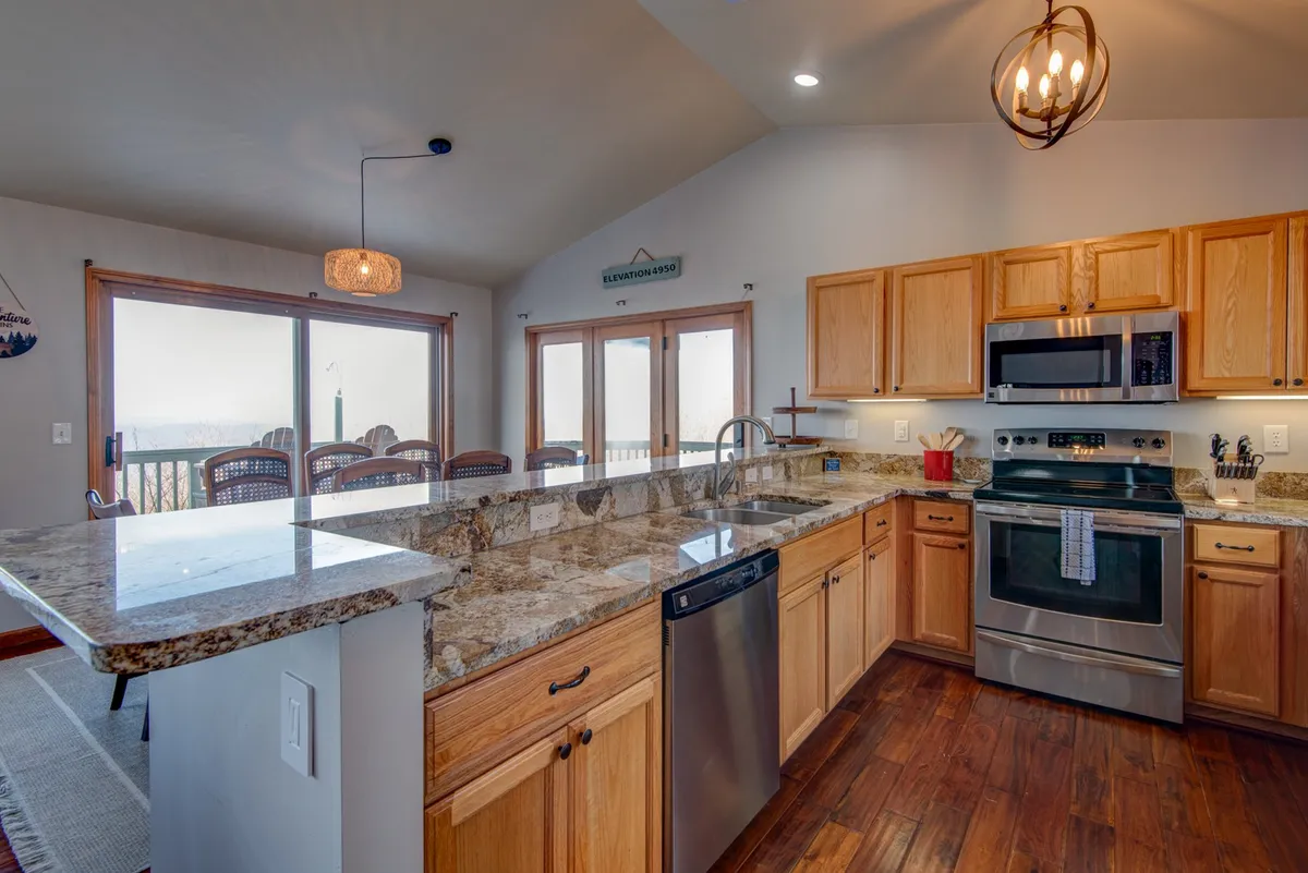 A bright, modern kitchen featuring granite countertops, stainless steel appliances, and warm wooden cabinetry. The kitchen seamlessly flows into a dining area with a scenic view visible through the window, emphasizing open-concept living and communal cooking and dining.