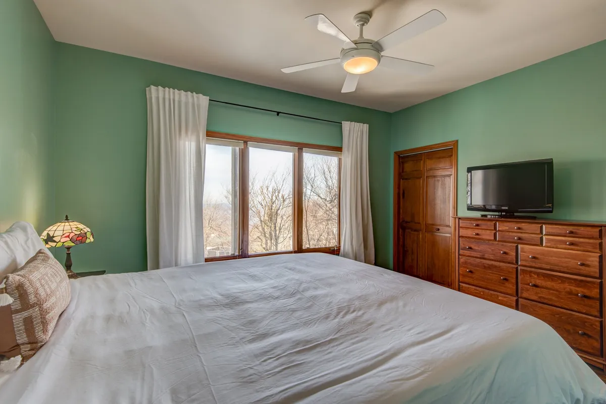 A bright, inviting bedroom with a large bed dressed in white linens, a bay window with multiple panes framing mountain views, a wooden dresser, a ceiling fan overhead, and a stained glass lamp adding a romantic touch. The window brings the outdoors inside and creates a tranquil atmosphere perfect for relaxation or enjoying peaceful mornings together.