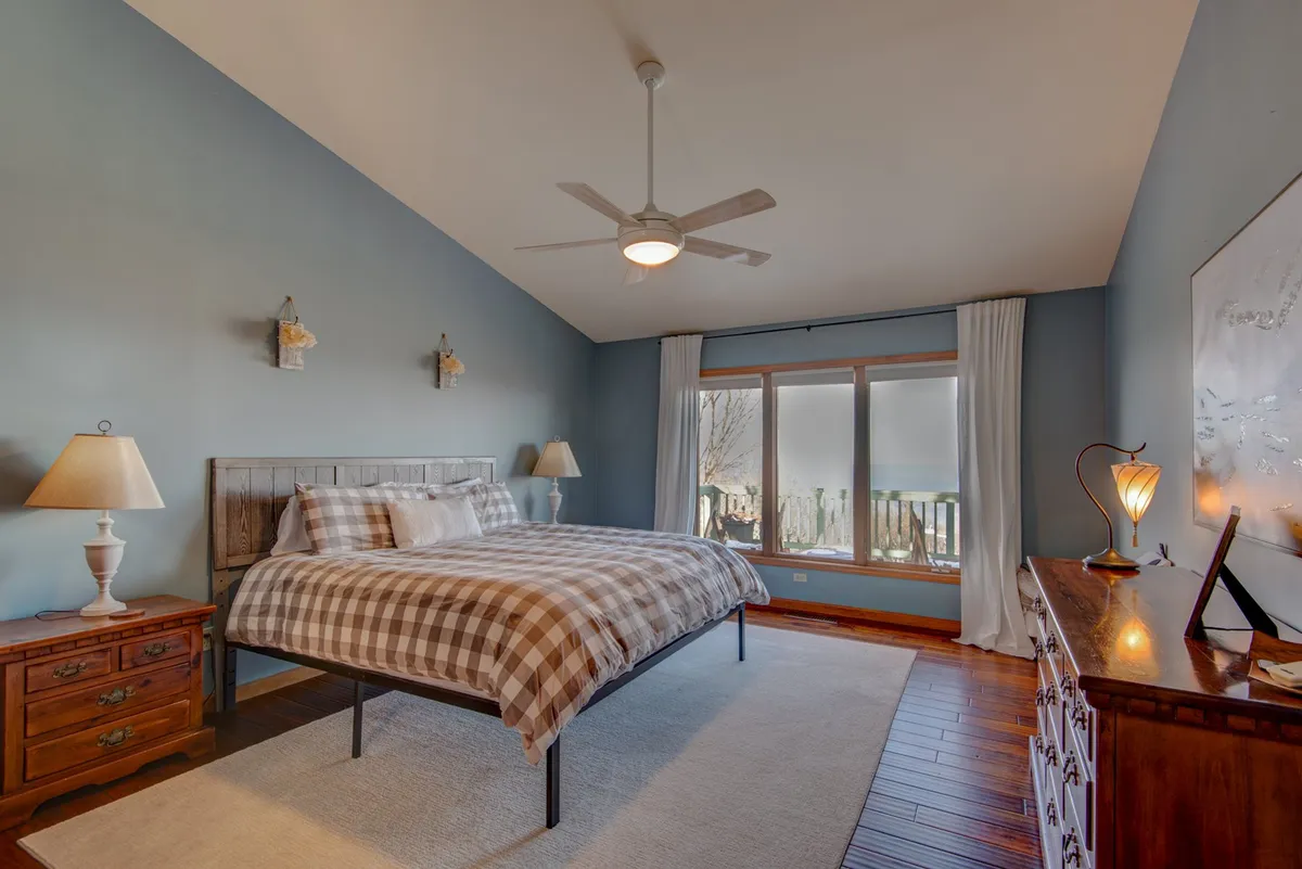 A sunlit bedroom furnished with a bed featuring a plaid bedspread, a large window that lets in natural light, a wooden dresser, and a nightstand with a lamp. The room exudes a calm, inviting feel with a touch of mountain charm.