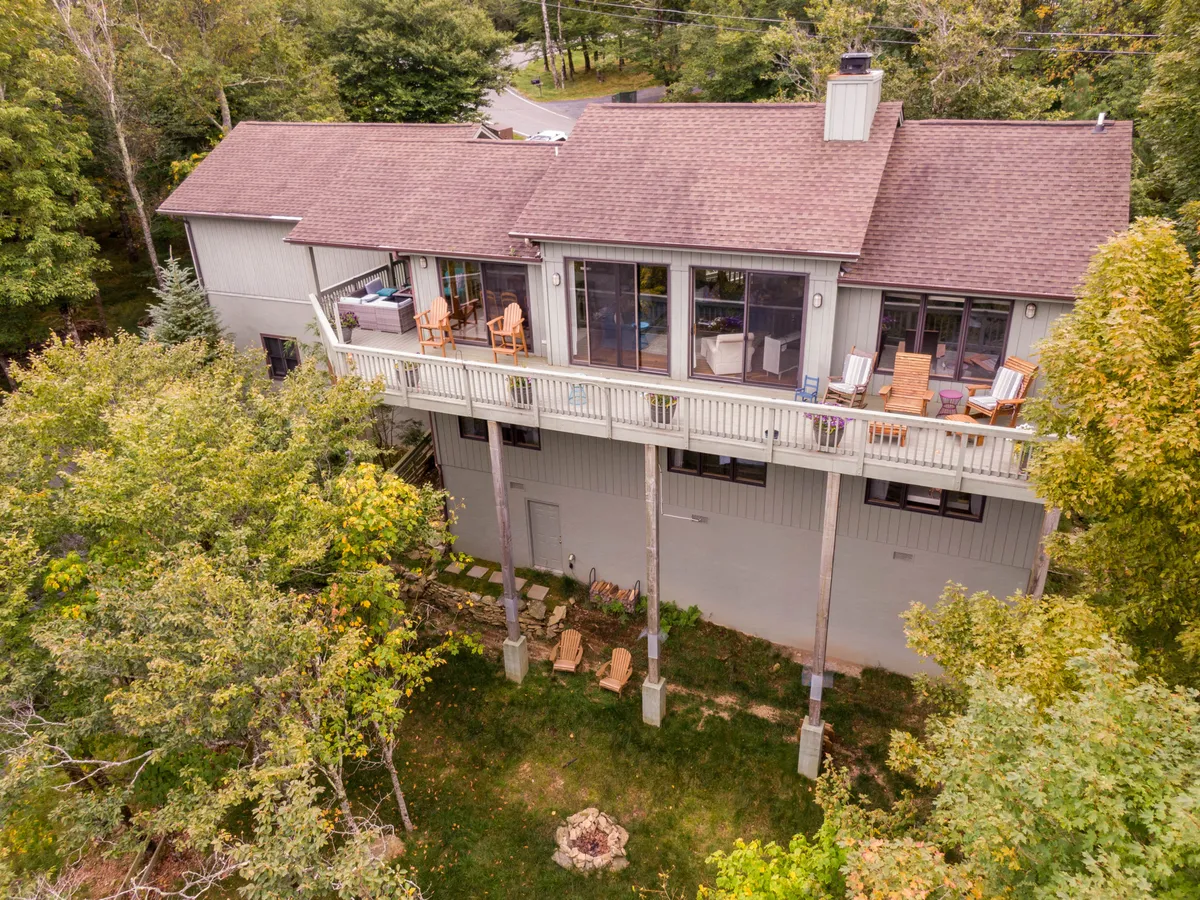 A wide shot of the elevated private deck featuring a comfortable seating area, large windows that blend indoor and outdoor living, and a fire pit below. The backdrop includes lush trees and mountain views, setting an inviting atmosphere for both daytime relaxation and evening gatherings.