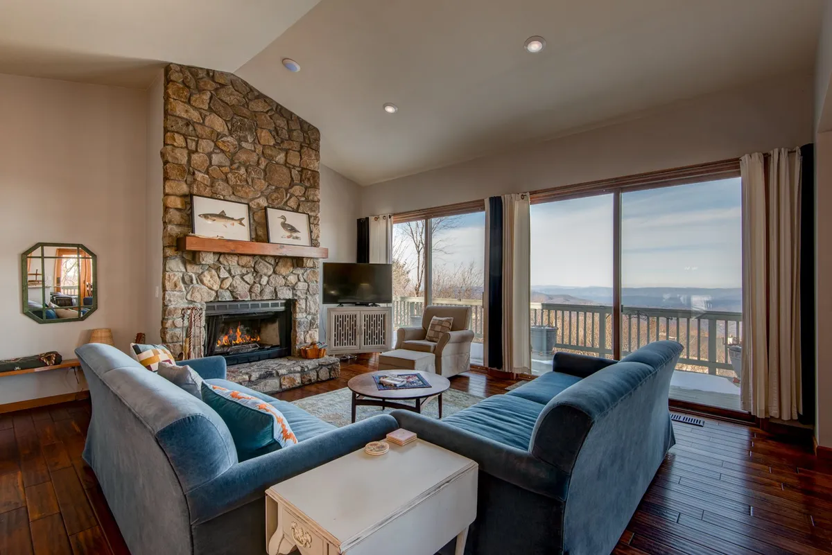 This image features a stone fireplace as the focal point, surrounded by a comfortable seating area with plush sofas. Towering windows fill the room with natural light and offer expansive, scenic views of Beech Mountain, perfectly capturing the cozy and inviting atmosphere of the living room. Guests are shown relaxing by the fire and enjoying the mountain scenery.
